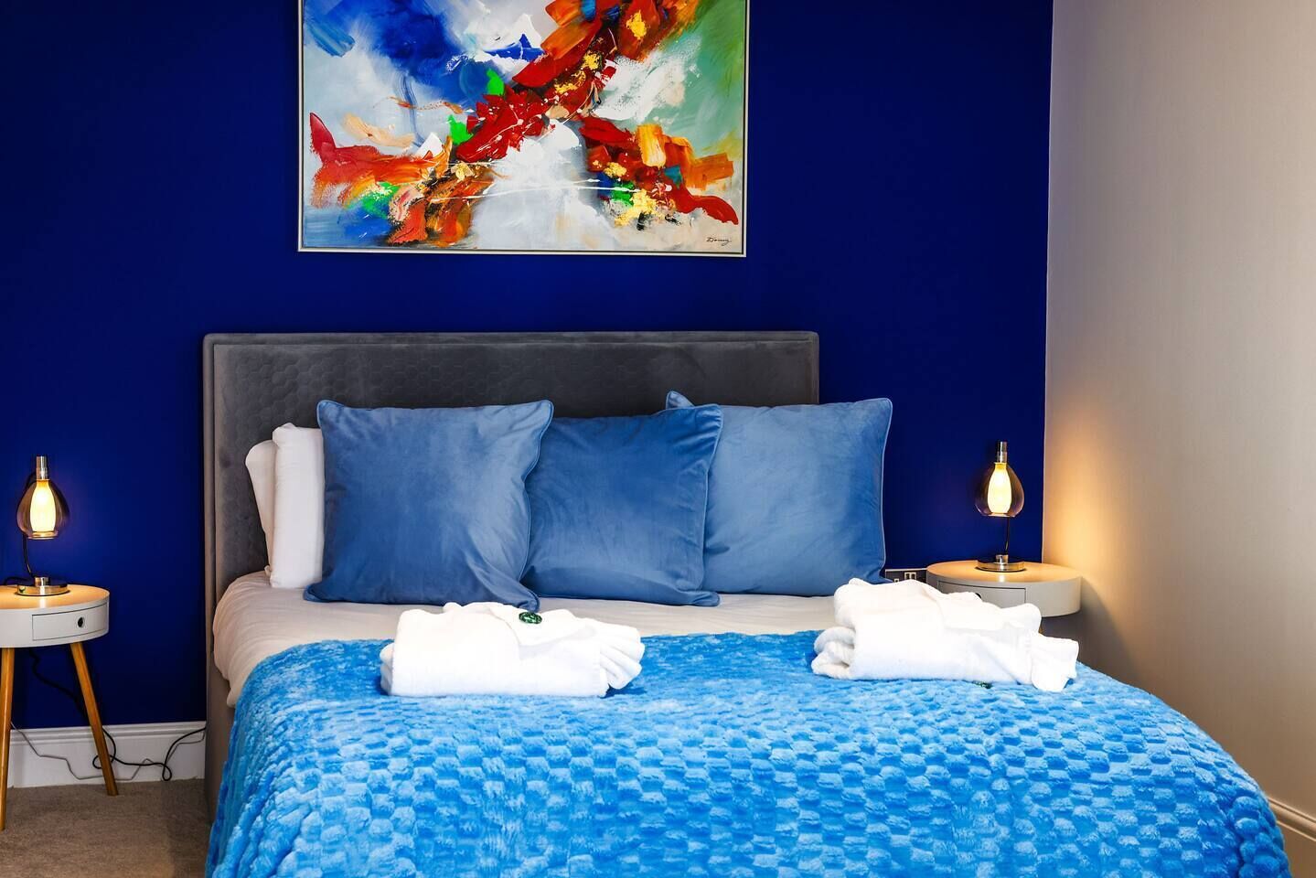 A bedroom with a blue wall, a large bed with blue bedding, two bedside lamps, and a colorful abstract painting above the bed.
