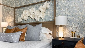 Ein ordentlich bezogenes Bett mit Kopfteil, ein Nachttisch mit Lampe und ein Blumenmuster an der Wand.