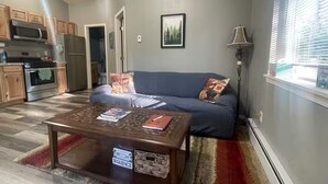 A living room with a sofa, a coffee table, a lamp, and a kitchen area with cabinets and appliances. - Adventure Stay Unit 2 (Palmer)