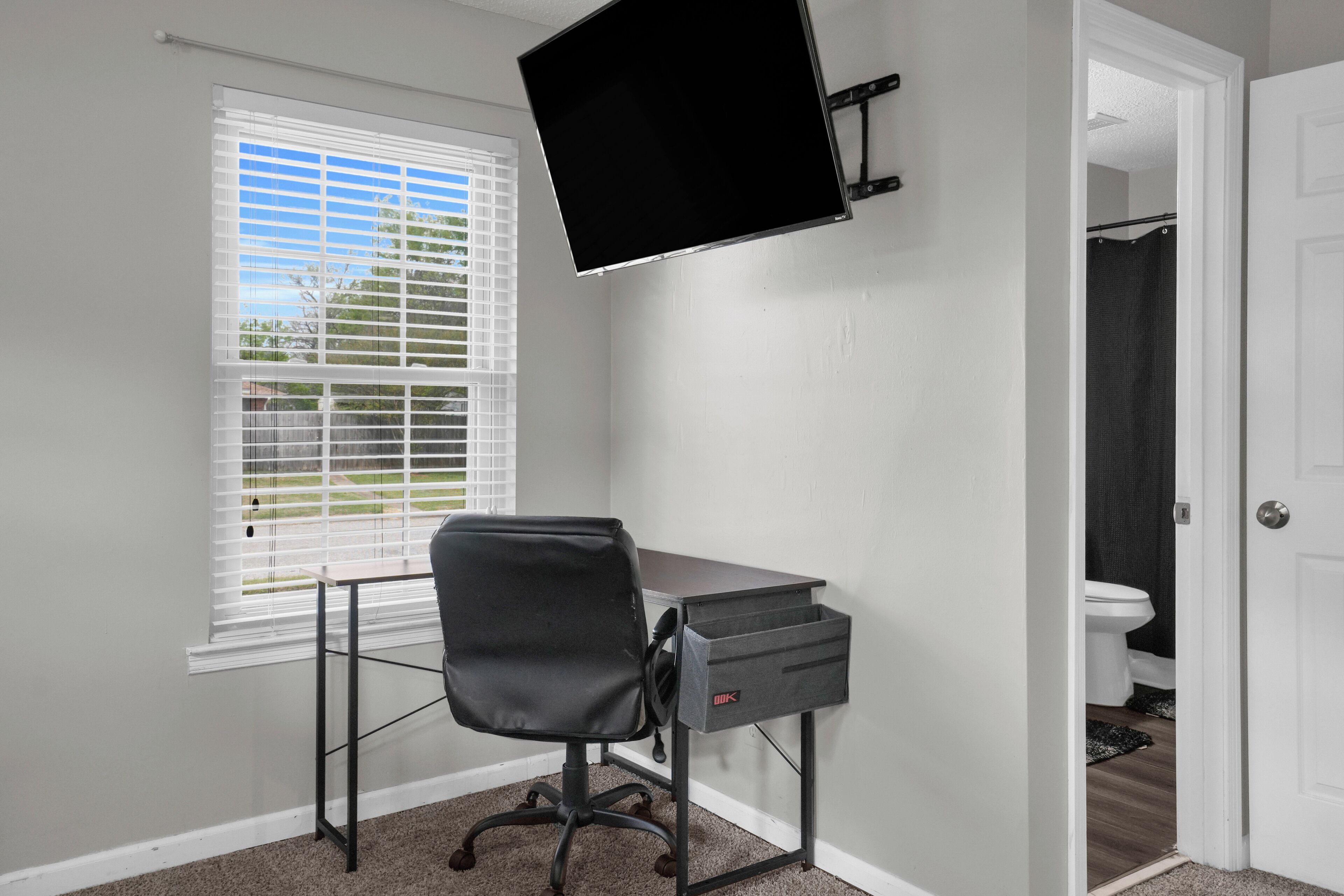 A room with a desk, chair, TV, window, and door leading to a bathroom.