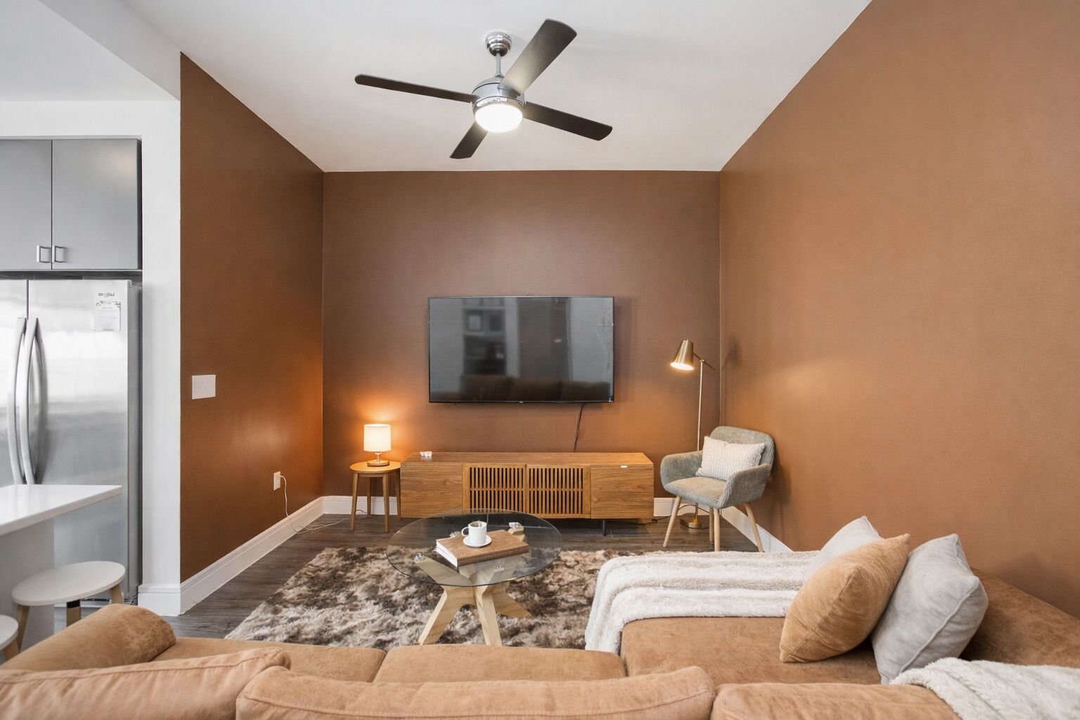 Een moderne woonkamer met een flatscreen-tv, een plafondventilator, een bank, een salontafel en een bijzettafeltje met een lamp.