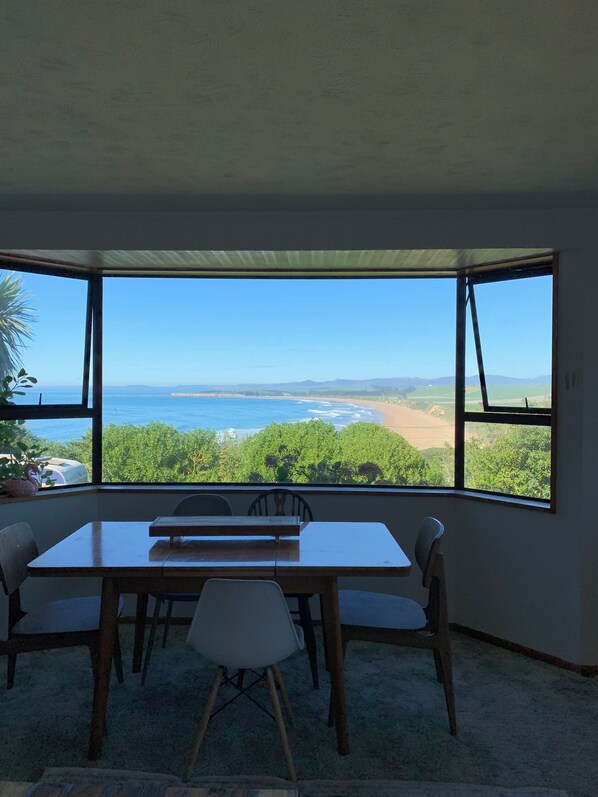 A room with a large window overlooking a beach and ocean, a wooden table with chairs, and a view of greenery.