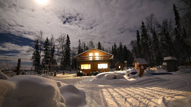 Uma cabana de madeira com varanda, rodeada por árvores cobertas de neve e um céu claro.