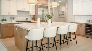 Una cucina moderna con mobili bianchi, un'isola centrale con sgabelli da bar, un frigorifero in acciaio inossidabile e illuminazione a sospensione.
