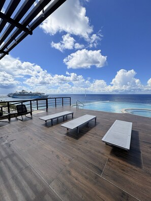 Une terrasse avec des chaises longues et une piscine, offrant une vue sur l’océan et un navire de croisière.