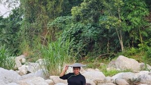 A person standing on rocks by a stream, another sitting on a rock, and a table with a plate. - Dong Que Homestay - Hostel (Ha Giang)