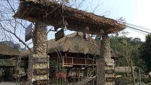 A rustic thatched-roof structure with stone pillars labeled with names, a wooden staircase, and a garden area. - Dong Que Homestay - Hostel (Ha Giang)