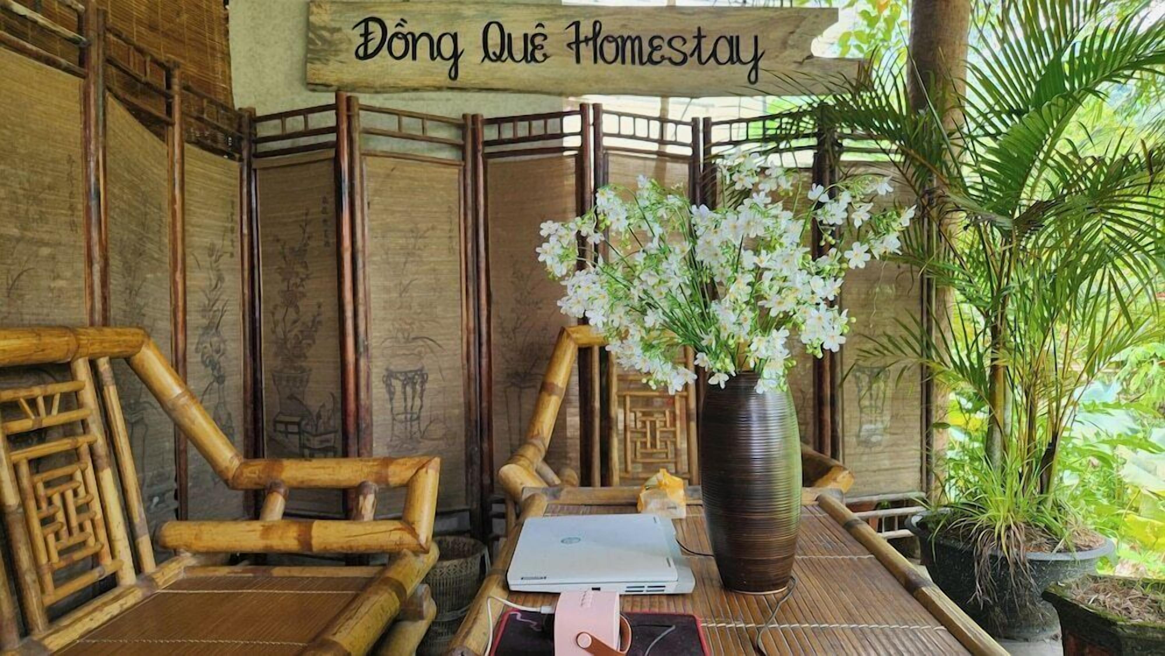 A bamboo-framed room with a laptop, a vase of flowers, and a sign reading 'Đồng Que Homestay'.