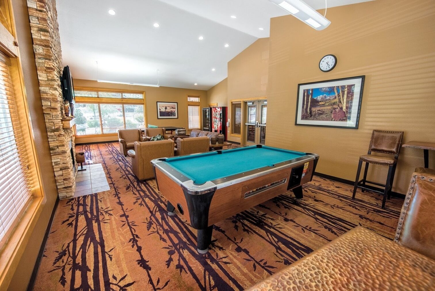 Une table de billard dans une pièce dotée d’un tapis à motifs, de chaises et d’une horloge au mur.