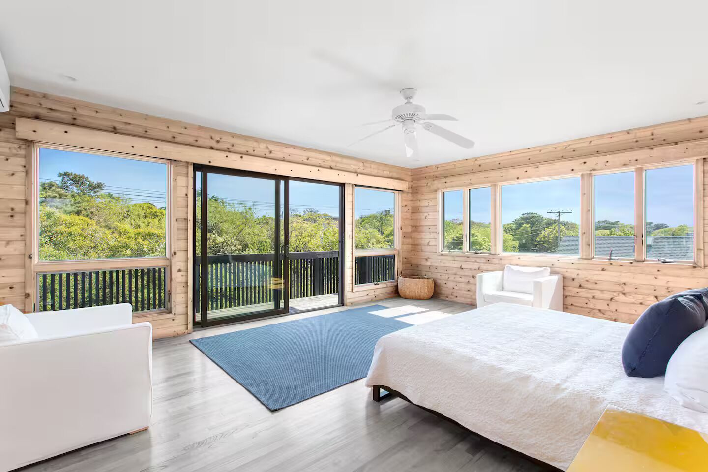 A modern bedroom with a large bed, a ceiling fan, and a view of greenery through large windows.