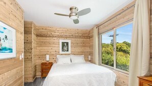 Quarto com cama, paredes de madeira, ventilador de teto, mesa de cabeceira, janela com vista e um quadro na parede.