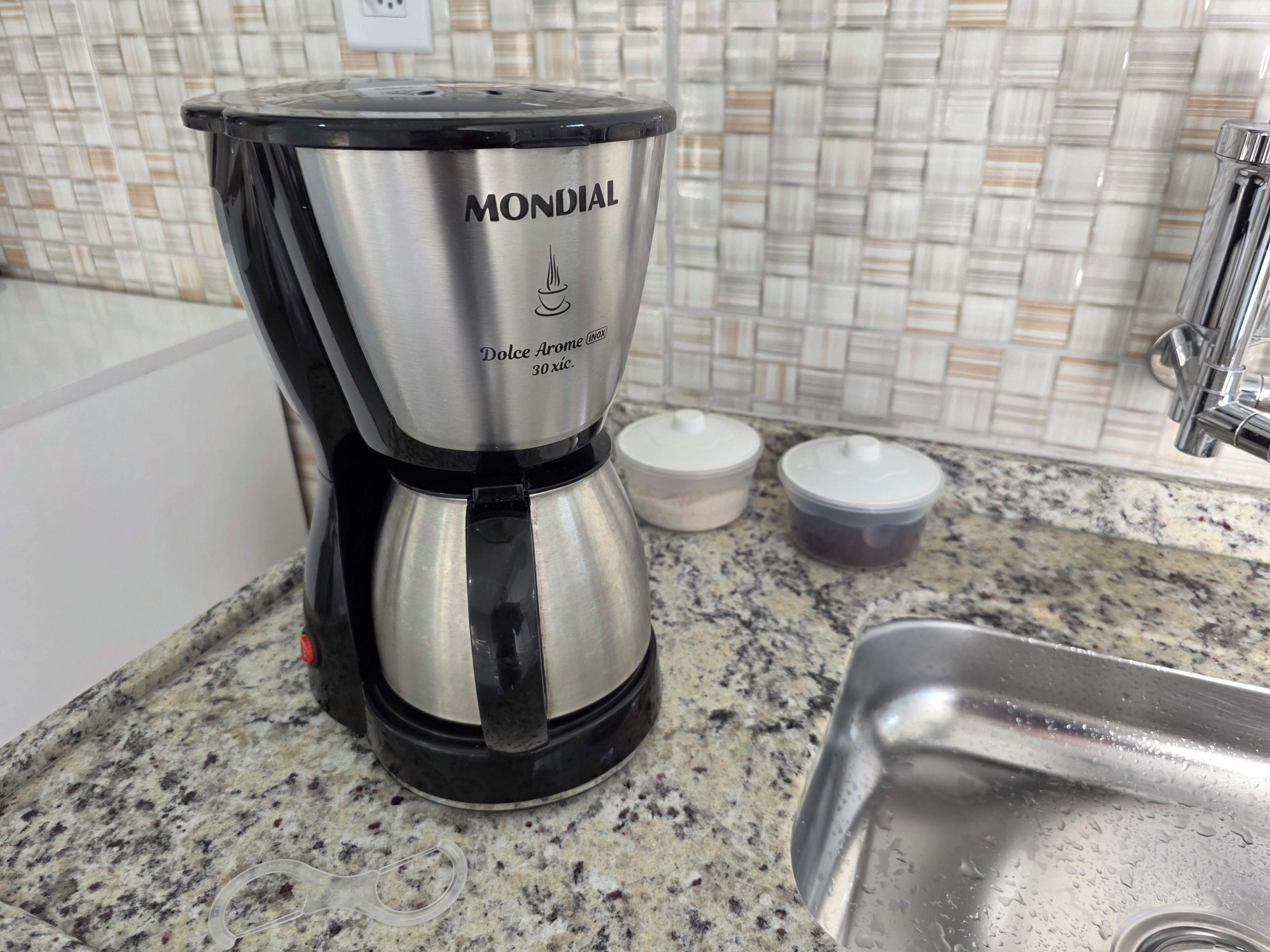 A stainless steel Mondial coffee maker on a granite countertop with a tiled backsplash and a stainless steel sink.
