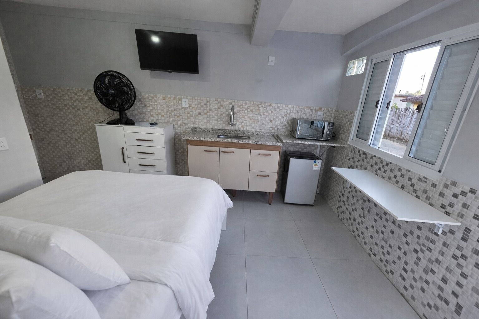 Quarto compacto com cama, pequeno espaço de cozinha, televisão e janela.