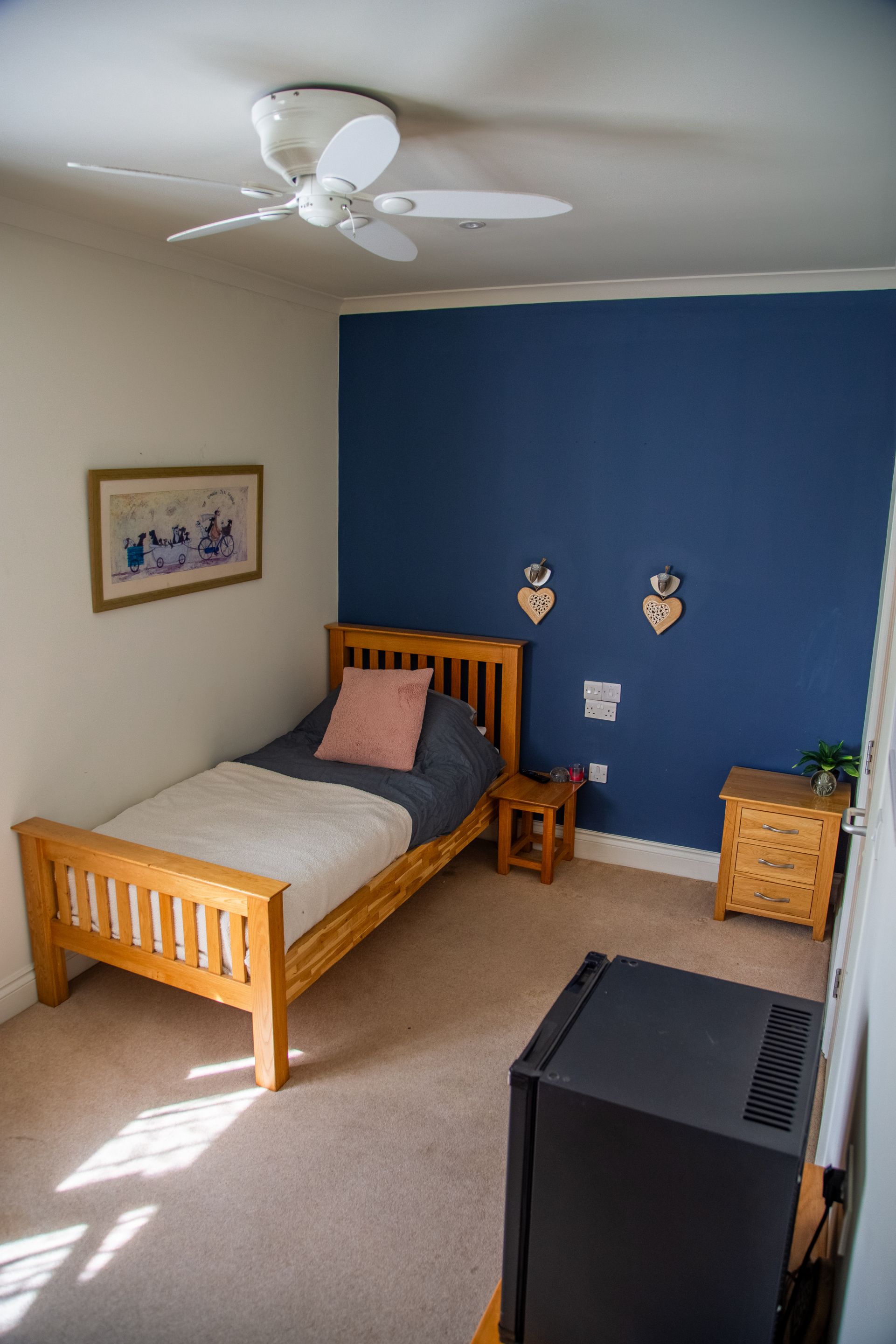 Un dormitorio con cama, una mesita de noche pequeña, un calefactor, televisor montado en la pared y un ventilador de techo.