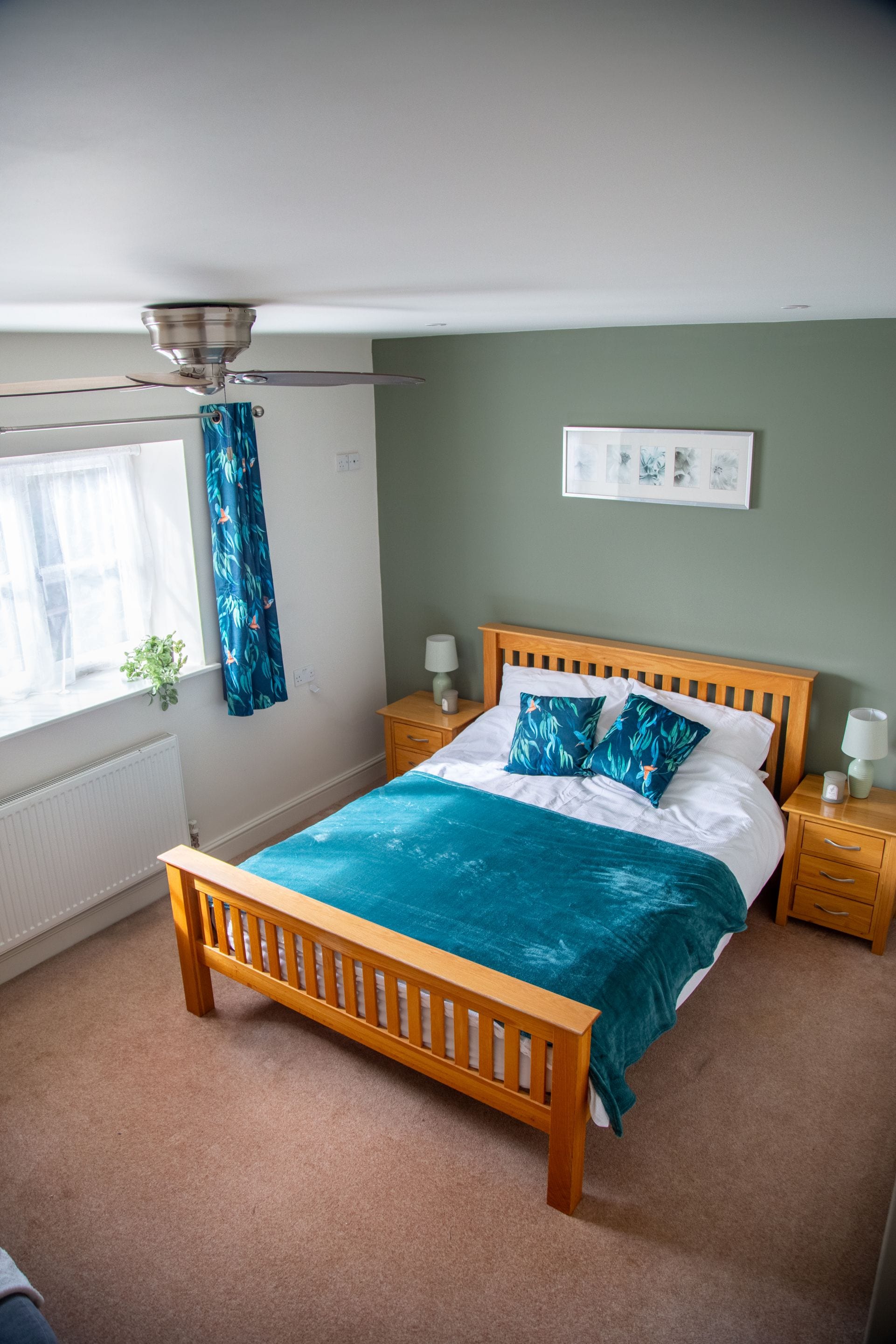 Een slaapkamer met een houten bed, een plafondventilator, een raam met gordijnen, een nachtkastje met een lamp en een aan de muur hangend schilderij.