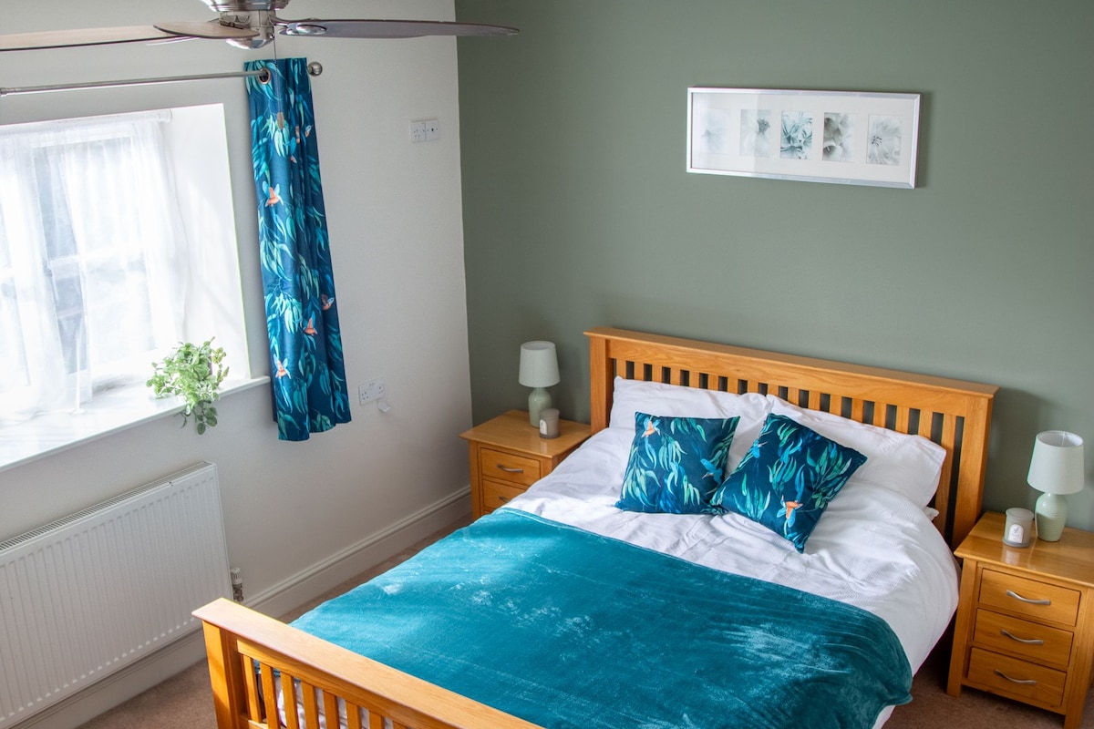 Un dormitorio con cama de madera, ventilador de techo, ventana con cortinas, mesita de noche con lámpara y un cuadro colgado en la pared.