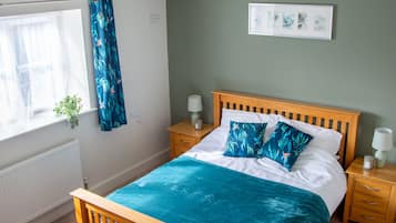 Een slaapkamer met een houten bed, een plafondventilator, een raam met gordijnen, een nachtkastje met een lamp en een aan de muur hangend schilderij.