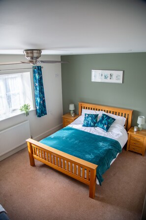 A bedroom with a wooden bed, a ceiling fan, a window with curtains, a bedside table with a lamp, and a wall-mounted picture.