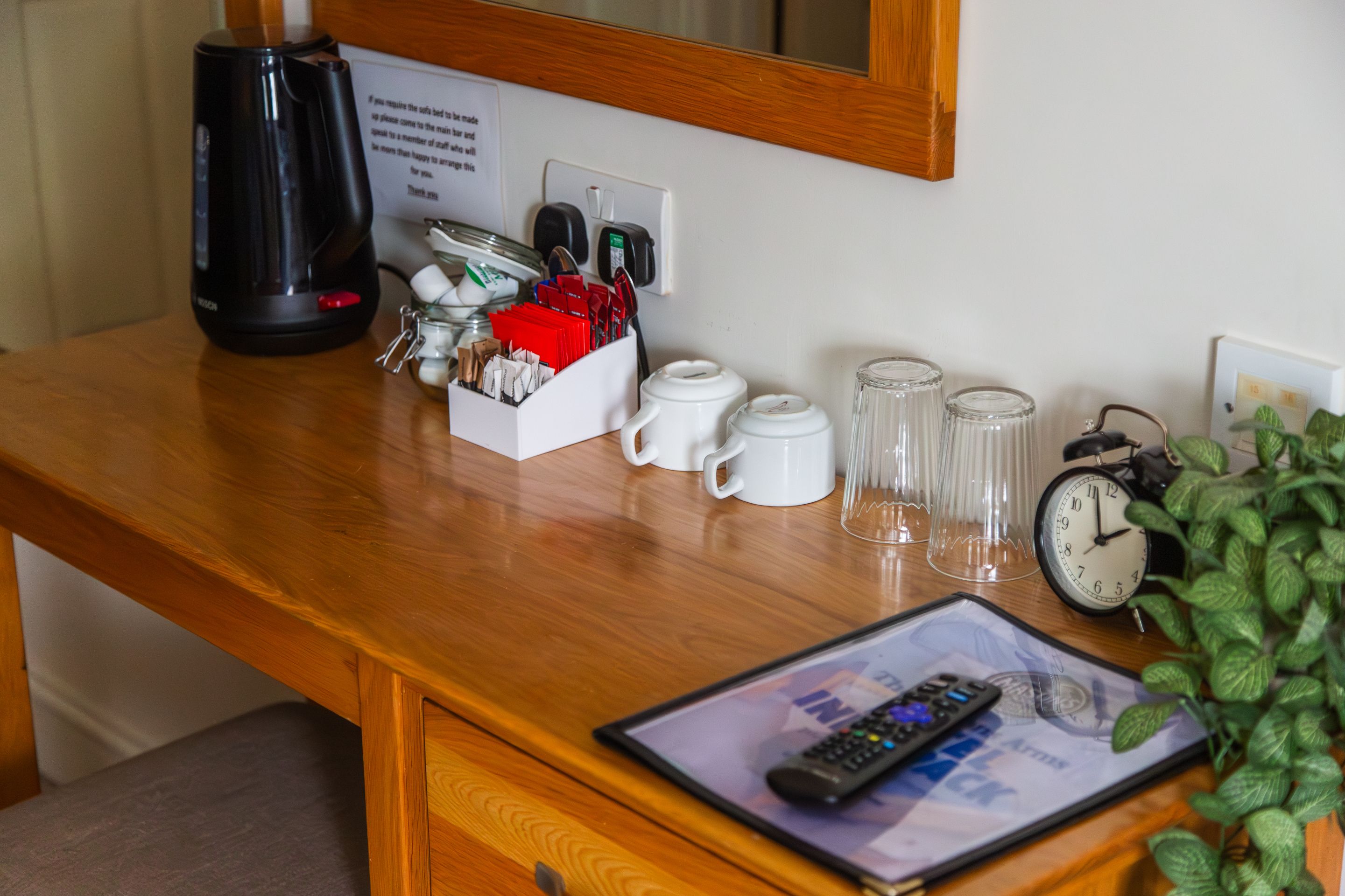 Escritorio de habitación de hotel con cafetera, porta taza, reloj y control remoto sobre una tablet.
