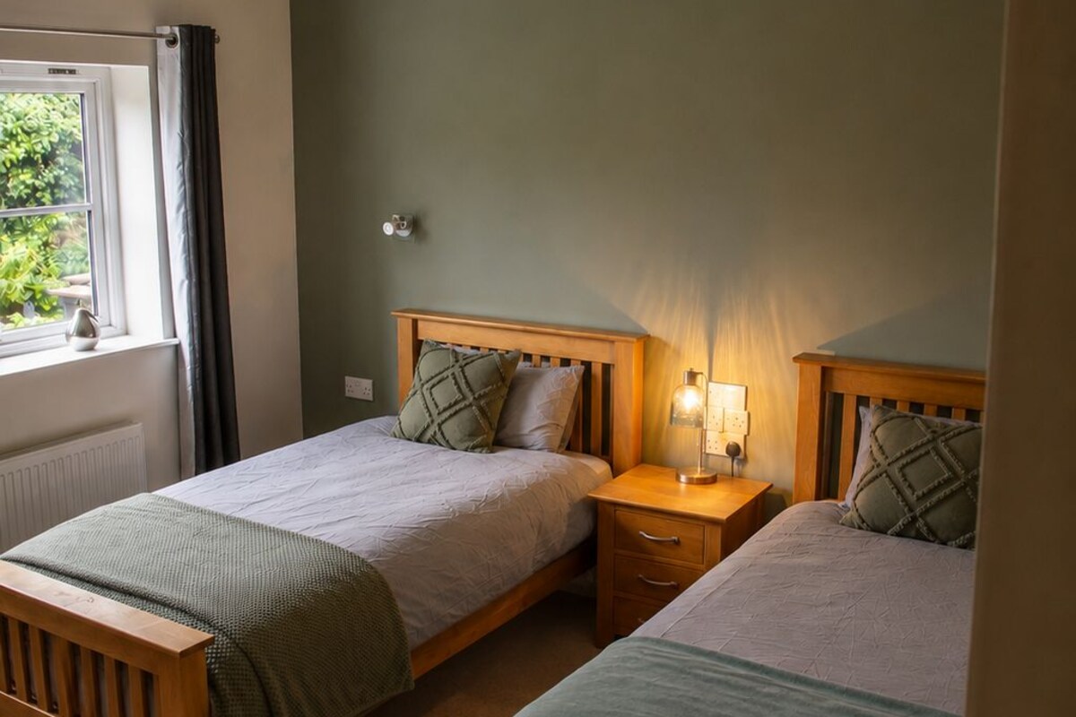 Un dormitorio con dos camas de madera, un ventilador de techo, una mesita de noche con lámpara y una ventana con cortinas.