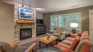 A living room with a stone fireplace, a television mounted on the wall, a sofa, a chair, and a window with blinds.