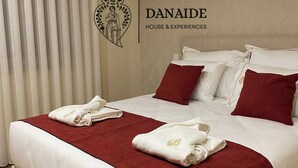 Una cama bien hecha con ropa de cama blanca y roja, un logotipo con forma de corazón en la pared y el texto 'DANAIDE HOUSE & EXPERIENCES'.