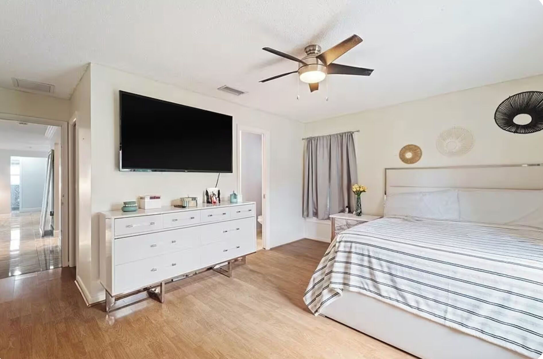 Un dormitorio con una cama, ventilador de techo, televisión y cómoda.
