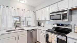 Una cocina moderna con gabinetes blancos, electrodomésticos de acero inoxidable y un ventanal con cortinas blancas.