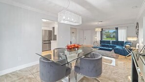 Una sala de estar moderna con una mesa de comedor de vidrio, sillas grises, un sofá azul y una araña.
