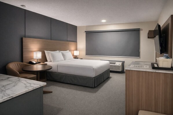 A modern hotel room with a bed, bedside table, lamp, chair, desk, and a flat-screen TV.