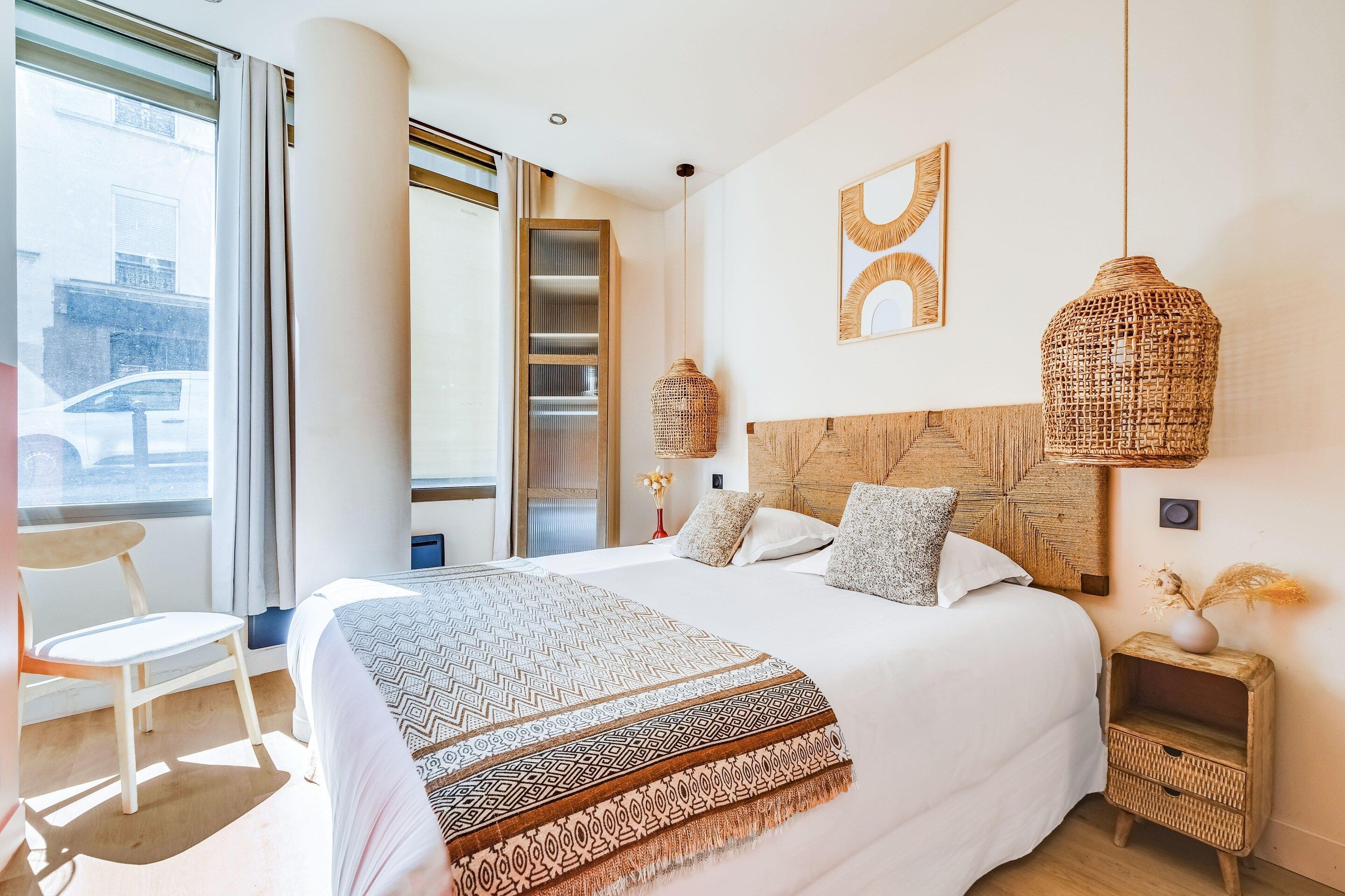 A hotel room with a large bed, a chair, a bedside table, a hanging light, and a view of the outside.