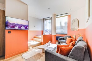 A modern living room with a flat-screen TV, a grey sofa with orange cushions, a white coffee table, and a staircase leading to an upper level. - Modern Apartment - 2bd/8p - Place D'italie (Paris)