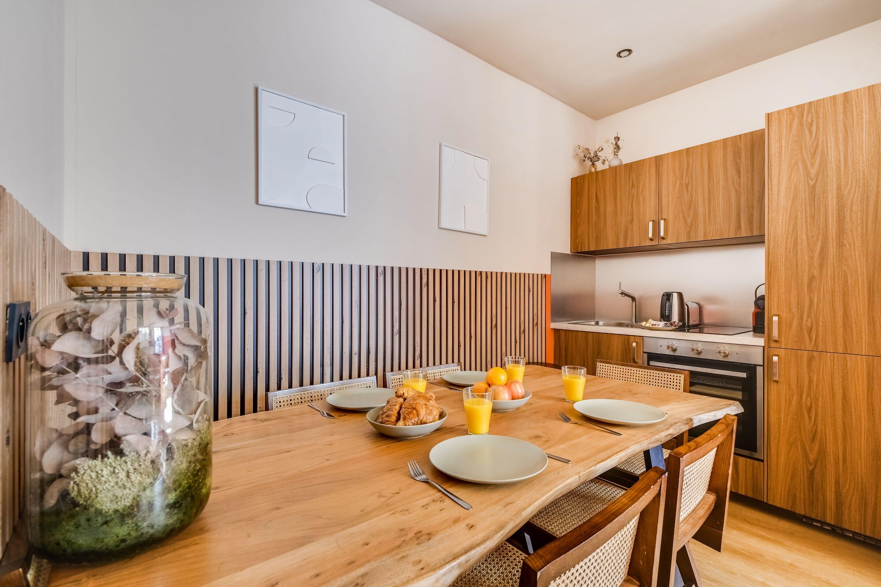 Eine moderne Küche mit Holzschränken, einem für das Frühstück gedeckten Esstisch und einem dekorativen Glas mit getrockneten Blumen.