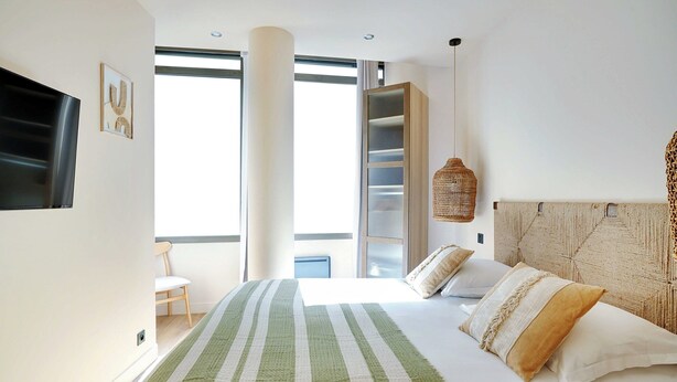 Un dormitorio moderno con cama, televisor, estantería y un amplio ventanal.