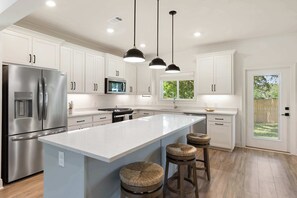 Una cocina moderna con armarios blancos, electrodomésticos de acero inoxidable, una isla grande con taburetes, iluminación colgante y vistas al exterior.