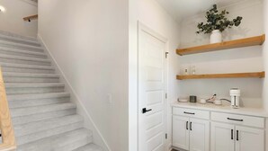 Una escalera con peldaños alfombrados, una puerta blanca, una zona de cocina con armarios blancos y estantes de madera, y una cafetera sobre la encimera.