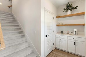 Eine Treppe mit Teppichstufen, eine weiße Tür, ein Küchenbereich mit weißen Schränken und Holzregalen sowie eine Kaffeemaschine auf der Arbeitsfläche.