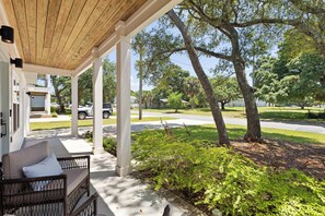A covered porch with a wicker chair and a cushion, a white building, and a street with trees and a parked car. - Spacious 3 Bedroom Home Near Beaches and Shopping! (Pensacola)