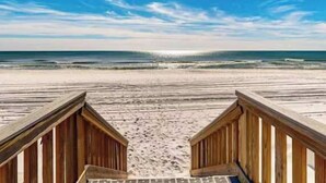 Ein hölzerner Steg führt zu einem Strand, im Hintergrund sind Wellen zu sehen.