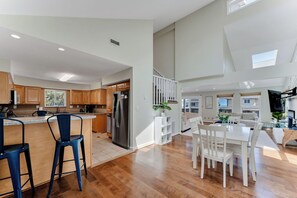 A spacious kitchen with wooden cabinets, a central island with bar stools, a dining area with a white table and chairs, and a staircase leading to an upper level. - Croatan Beach Spacious 4BR Home with Views (Virginia Beach)