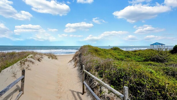 Ein Sandweg am Strand führt zum Meer, begleitet von einem Holenzaun und einer blauen Struktur in der Ferne.