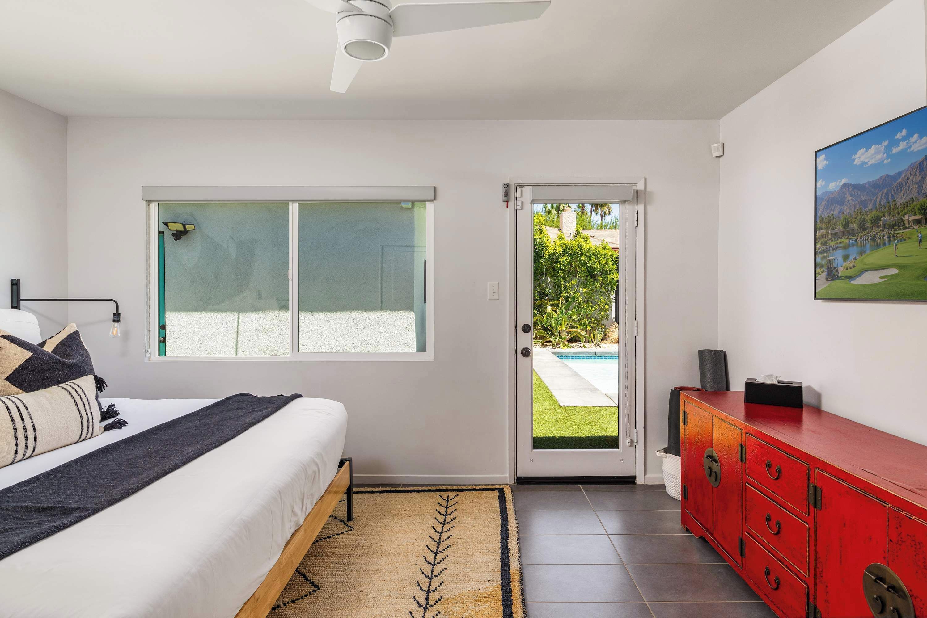Een slaapkamer met een bed, een raam, een deur naar buiten, een rode dressoir en een schilderij aan de muur.