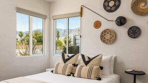 A bedroom with a bed, pillows, a bedside table, a lamp, and wall decorations. - Palm Springs Mid-Century Home | Pool, Spa, Casita + Sweeping Mountain Views (Palm Springs)