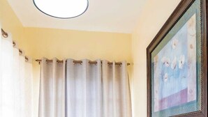 Un comedor con una mesa de vidrio, dos sillas, una lámpara de techo, una cortina y un cuadro en la pared.