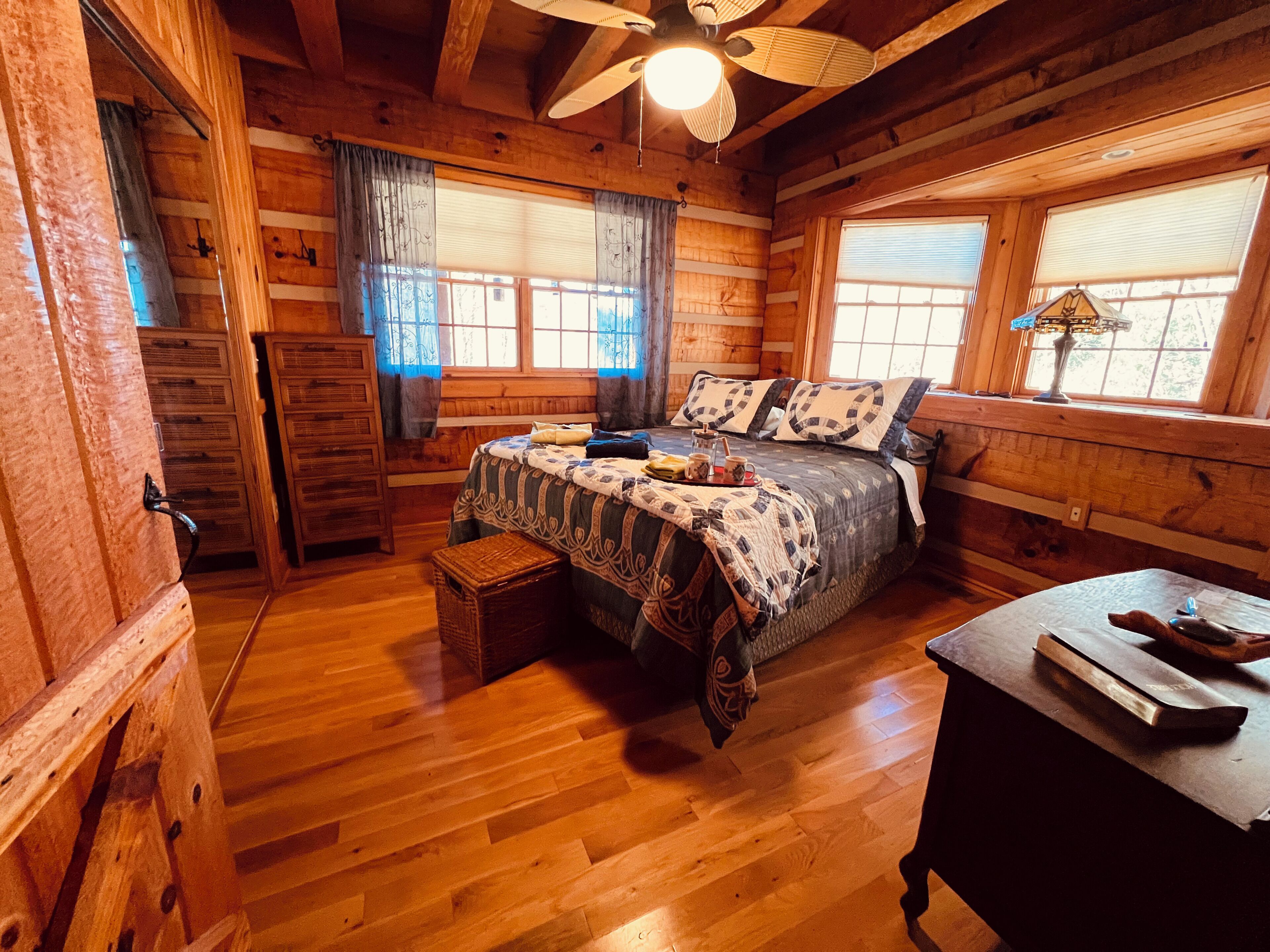A wooden cabin bedroom with a bed, a chest of drawers, a window with curtains, and a ceiling fan.