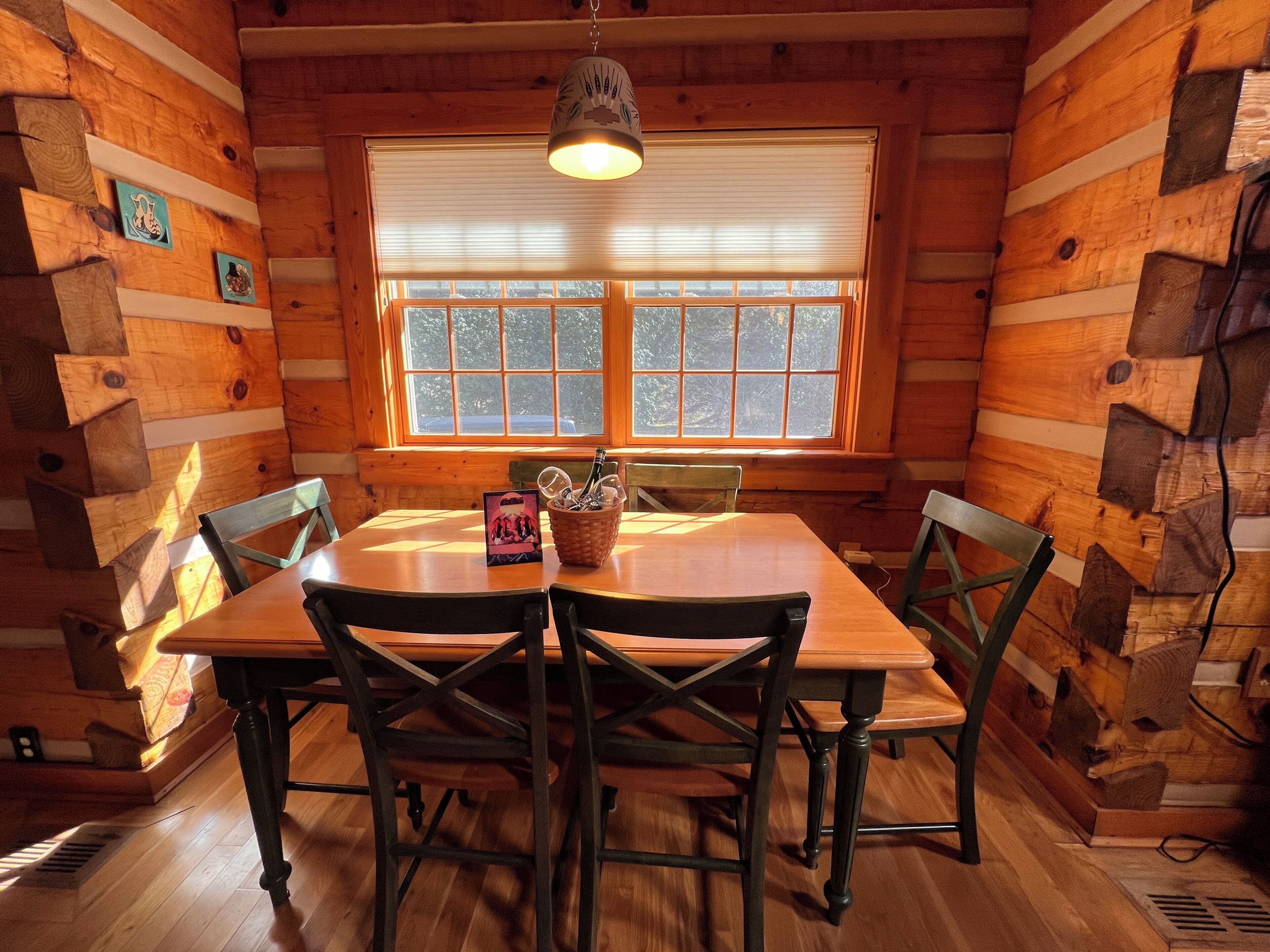 A wooden cabin interior with a dining table, four chairs, a hanging light fixture, and a window with blinds.