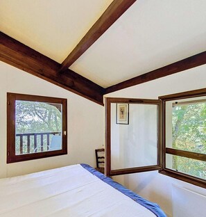 Ein Schlafzimmer mit einem Bett, zwei Fenstern, einer Holzbalkendecke und Blick auf Bäume.