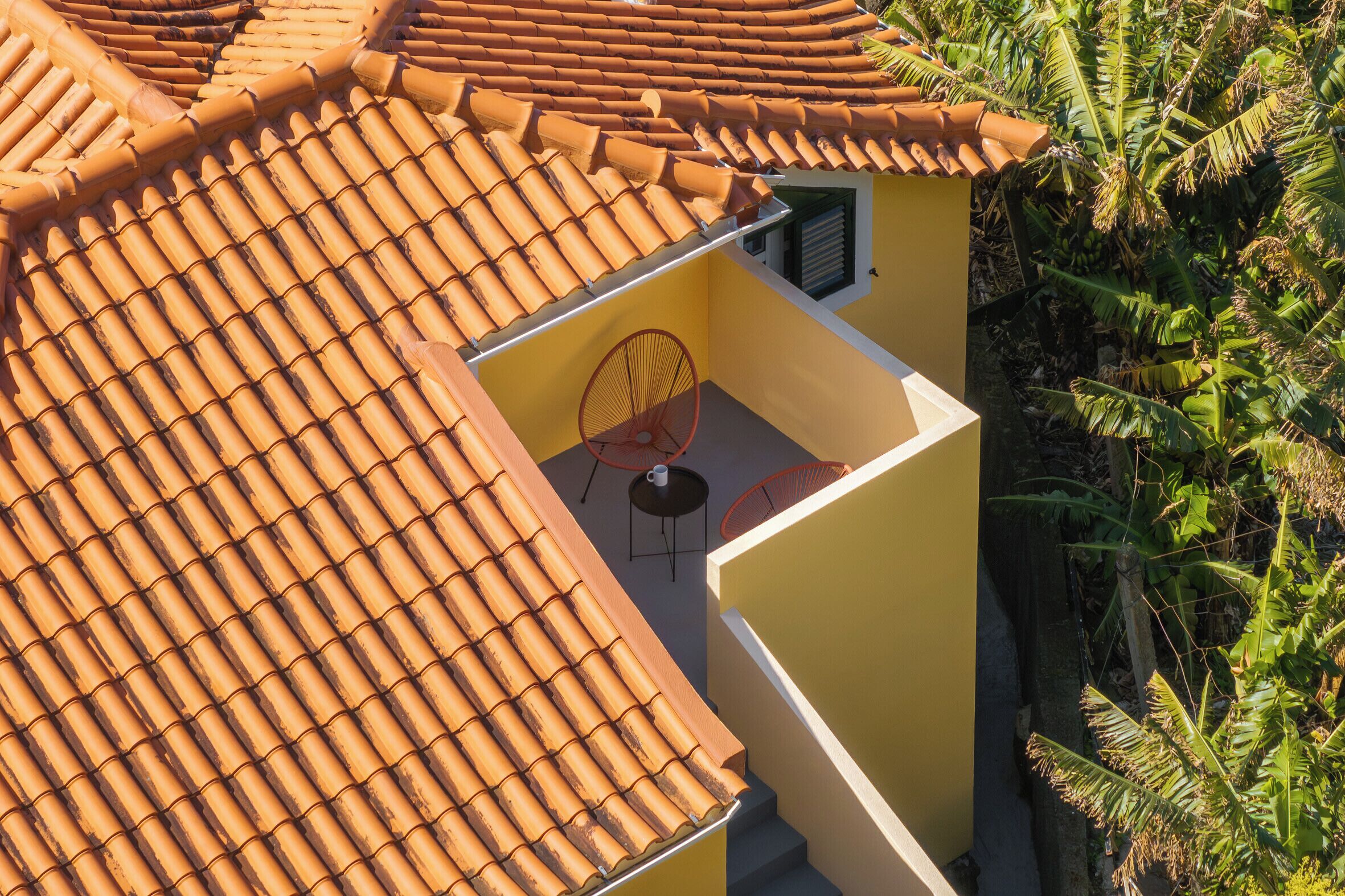 Ein gelbes Gebäude mit einem Ziegeldach, einem kleinen Balkon mit einem Stuhl und einem Fenster mit Rollläden.