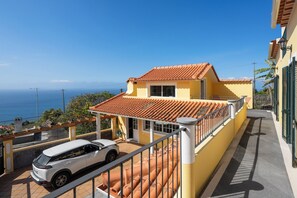A yellow building with a red-tiled roof, a white car parked in front, and a clear view of the sea.