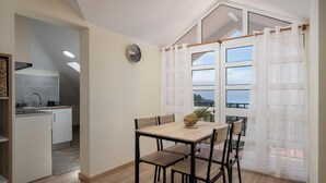 Ein Essbereich mit Tisch und Stühlen, eine Küche im Hintergrund und ein Ausblick durch ein Fenster mit Vorhängen.
